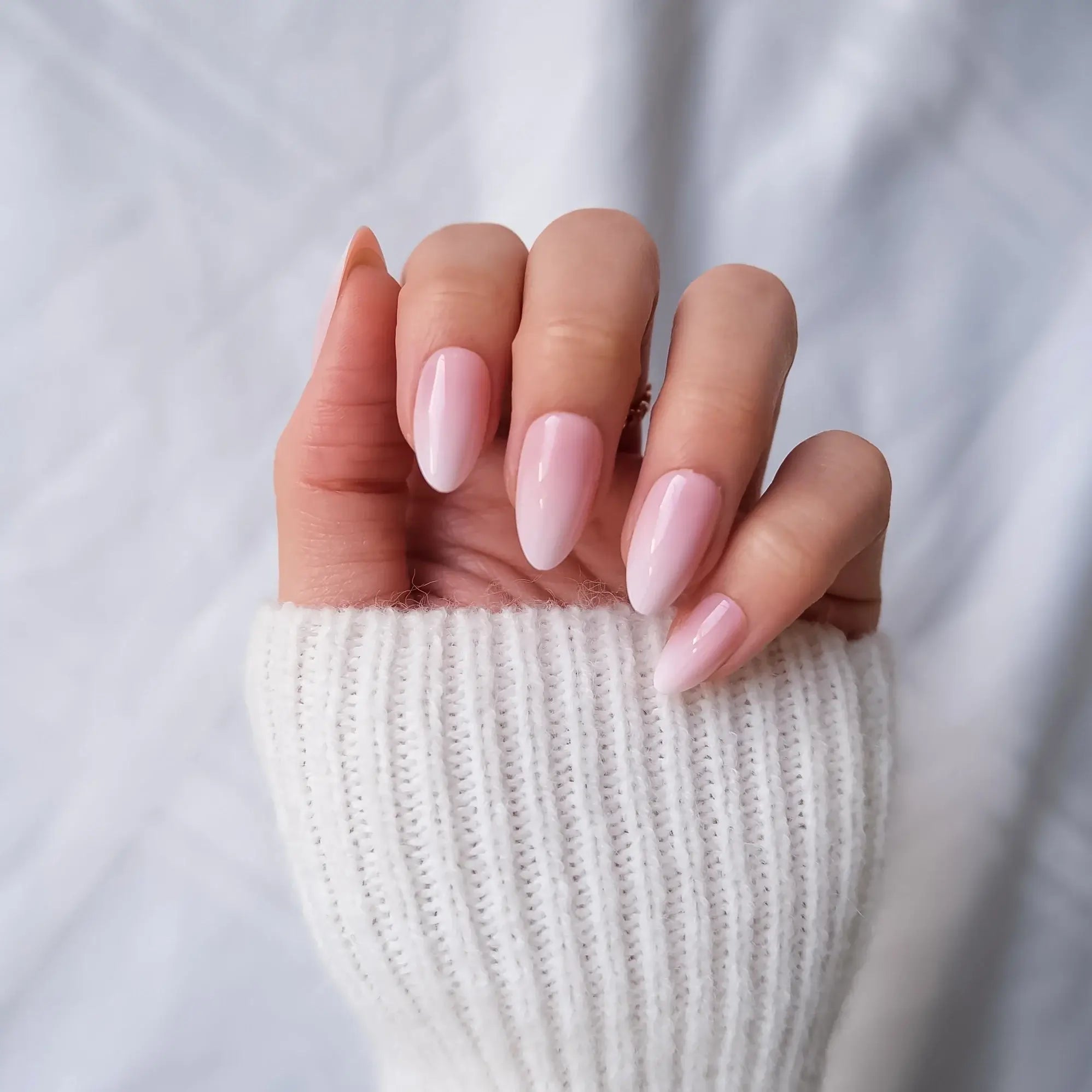 Hand with pink to white gradient press-on nails behind a white textured fabric