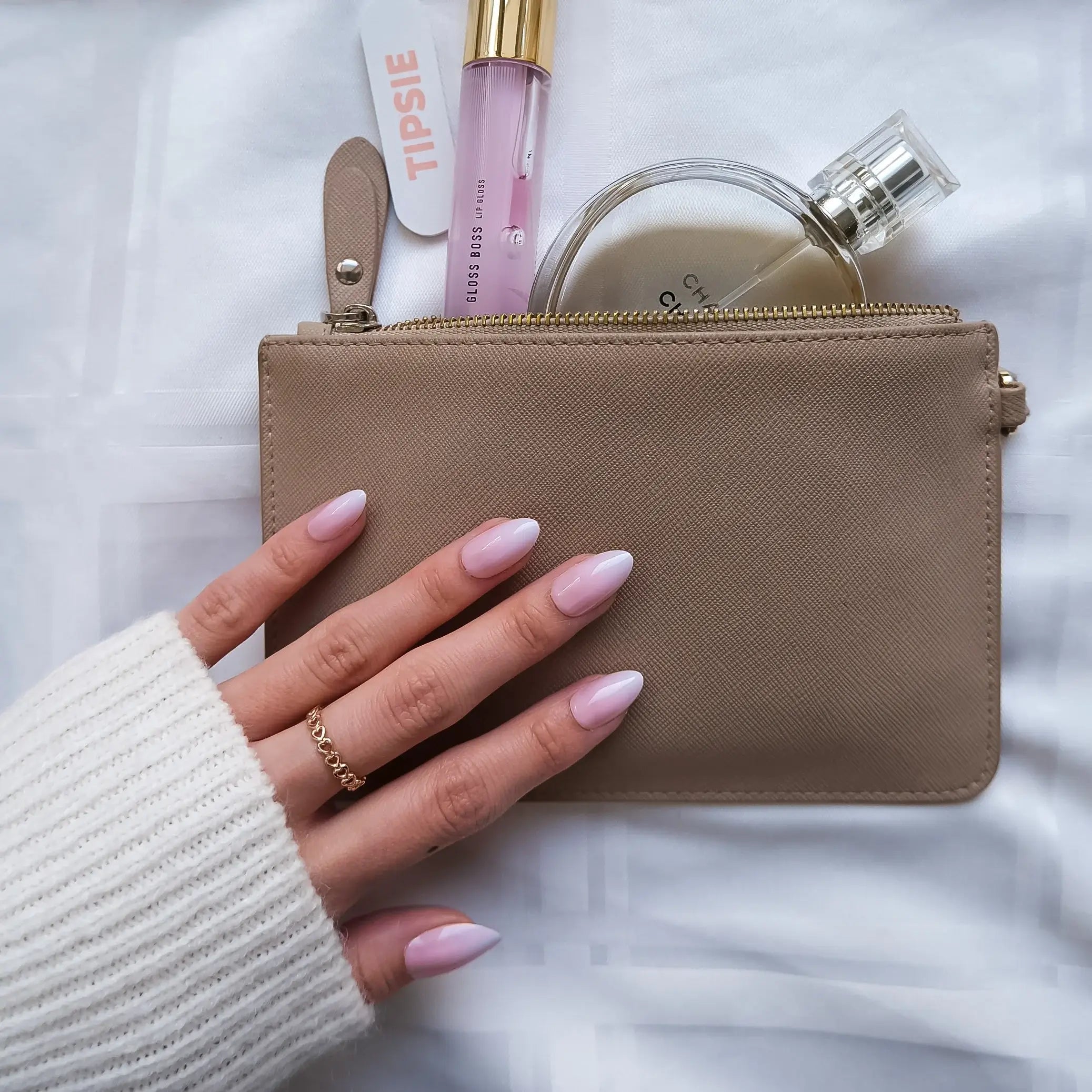 Beige pouch with lip gloss and perfume bottle held by a hand with pink to white gradient press on nail polish on a white background