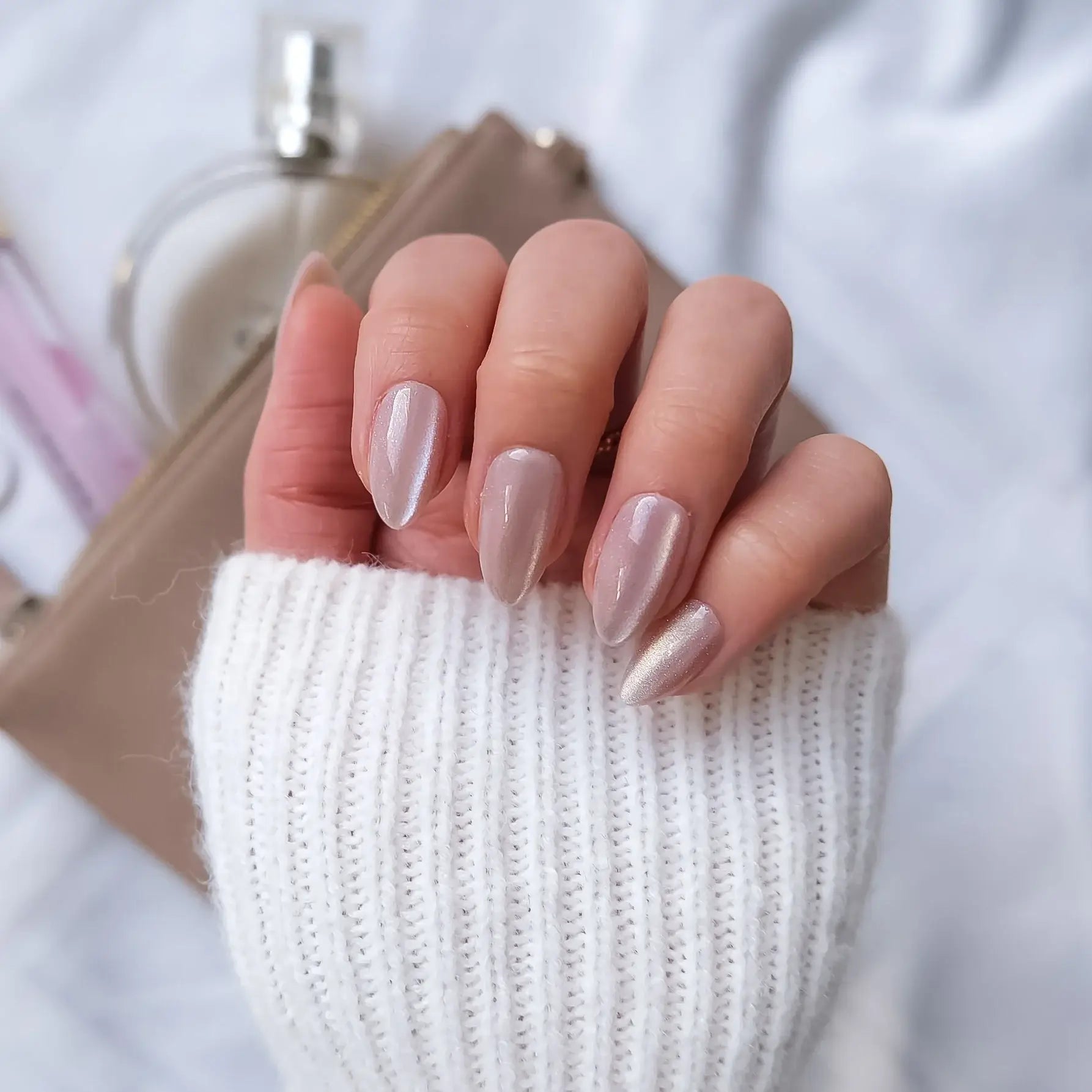 Close-up of a hand with subtle pink sparkly nail polish holding a brown clutch, with a white sweater and blurred perfume bottle in the background.