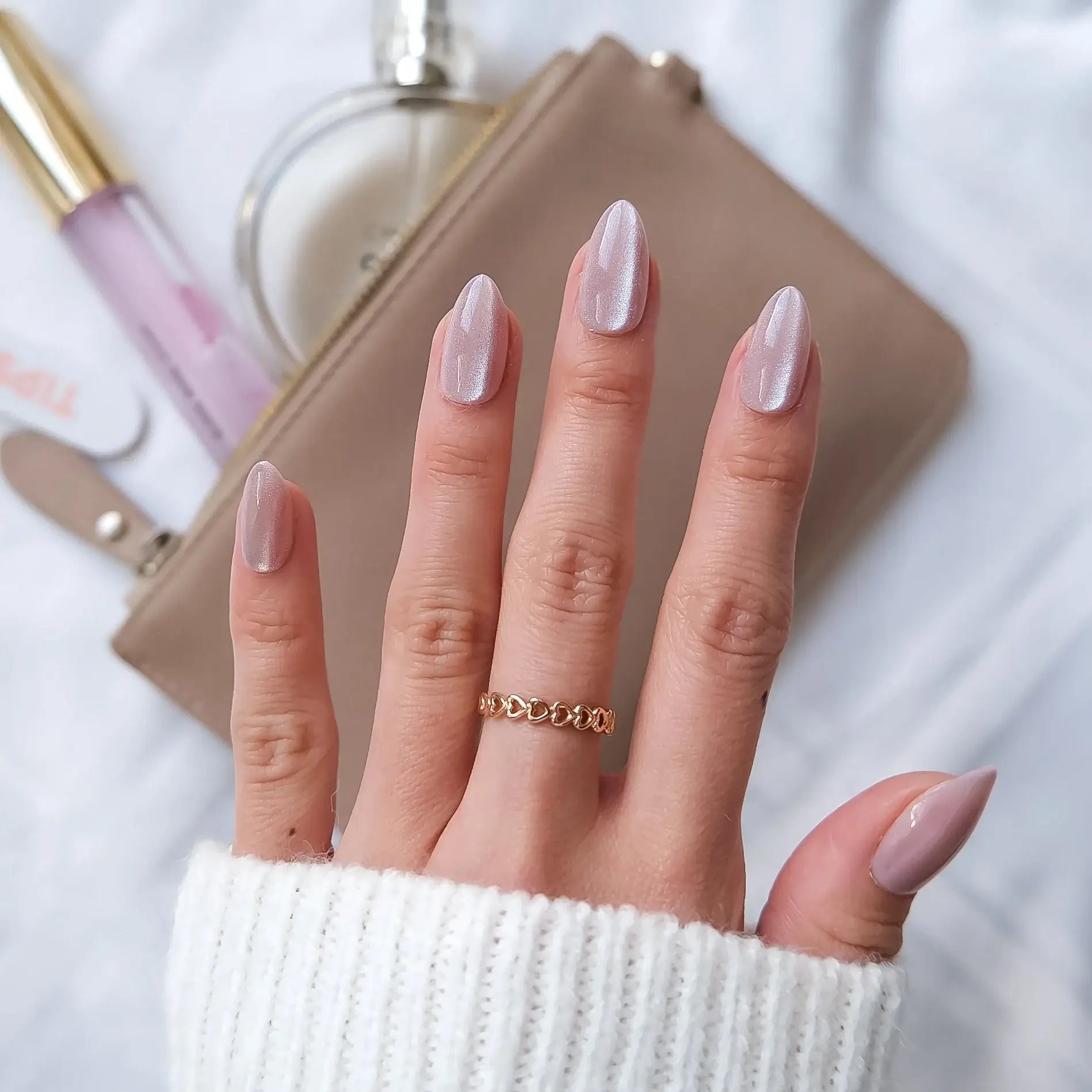 Hand with subtle pink sparkly press-on nails wearing a gold ring, with makeup items in the background.