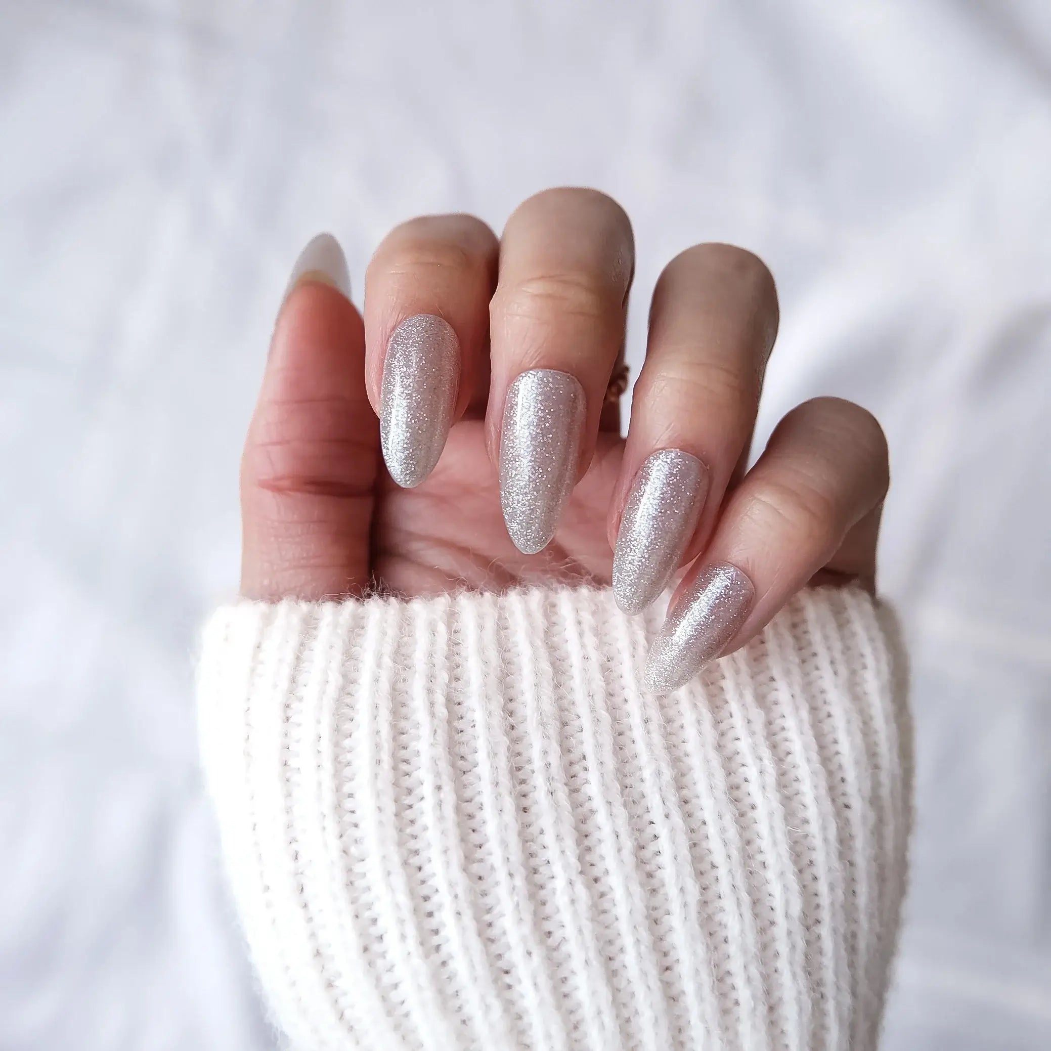Hand with glittery silver nail polish press on nails wearing a white knit sleeve against a light background