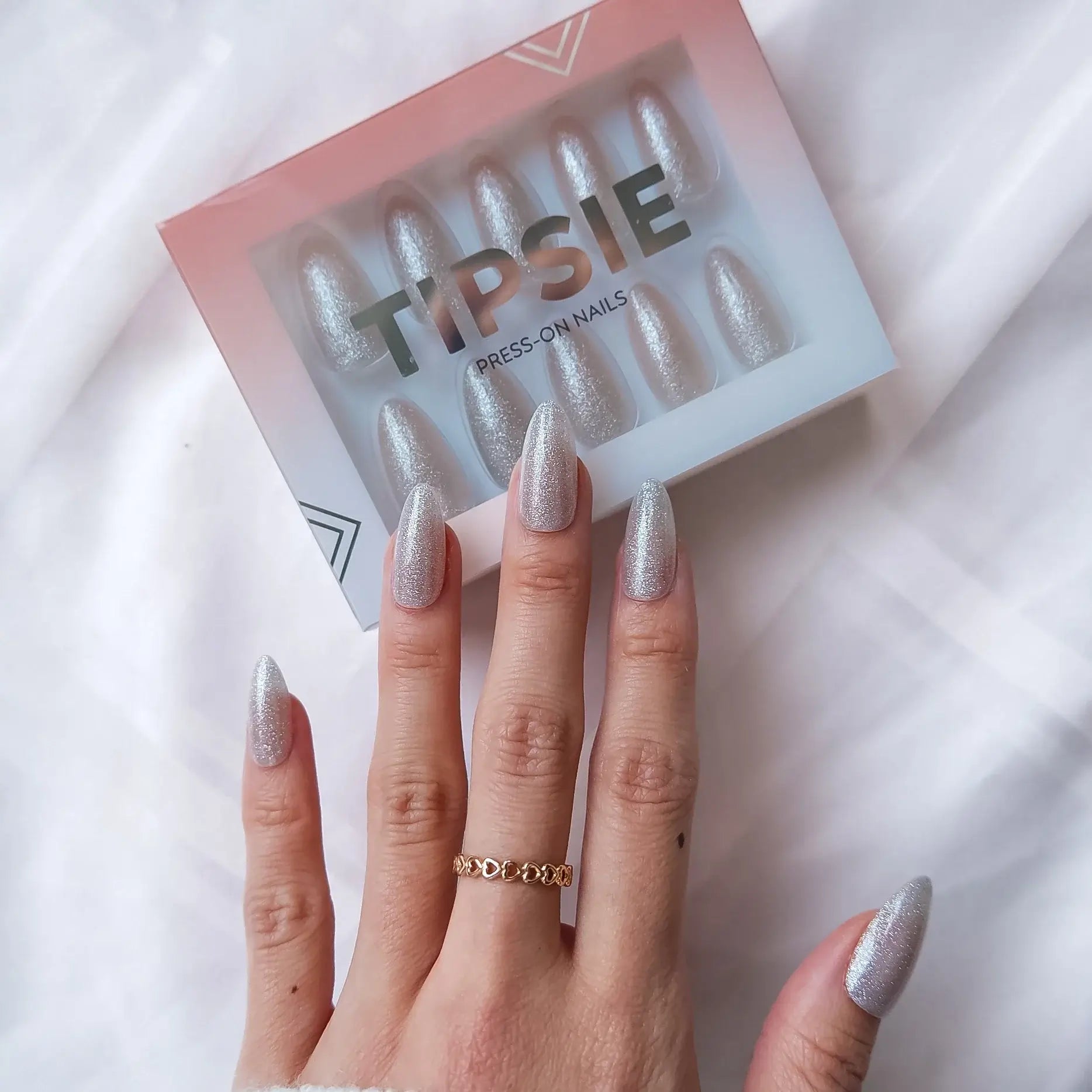 Hand with glittery press-on nails wearing a gold ring, next to a 'Tipsie' press-on nail box on a white background