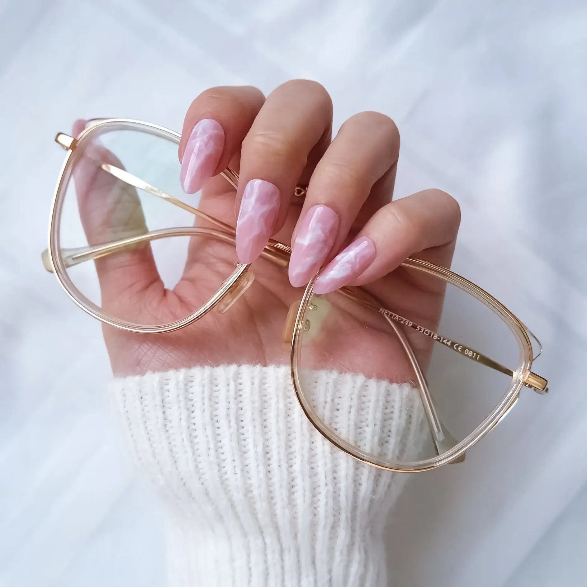 Gold-framed eyeglasses held by a hand with pink press on nails on a white background