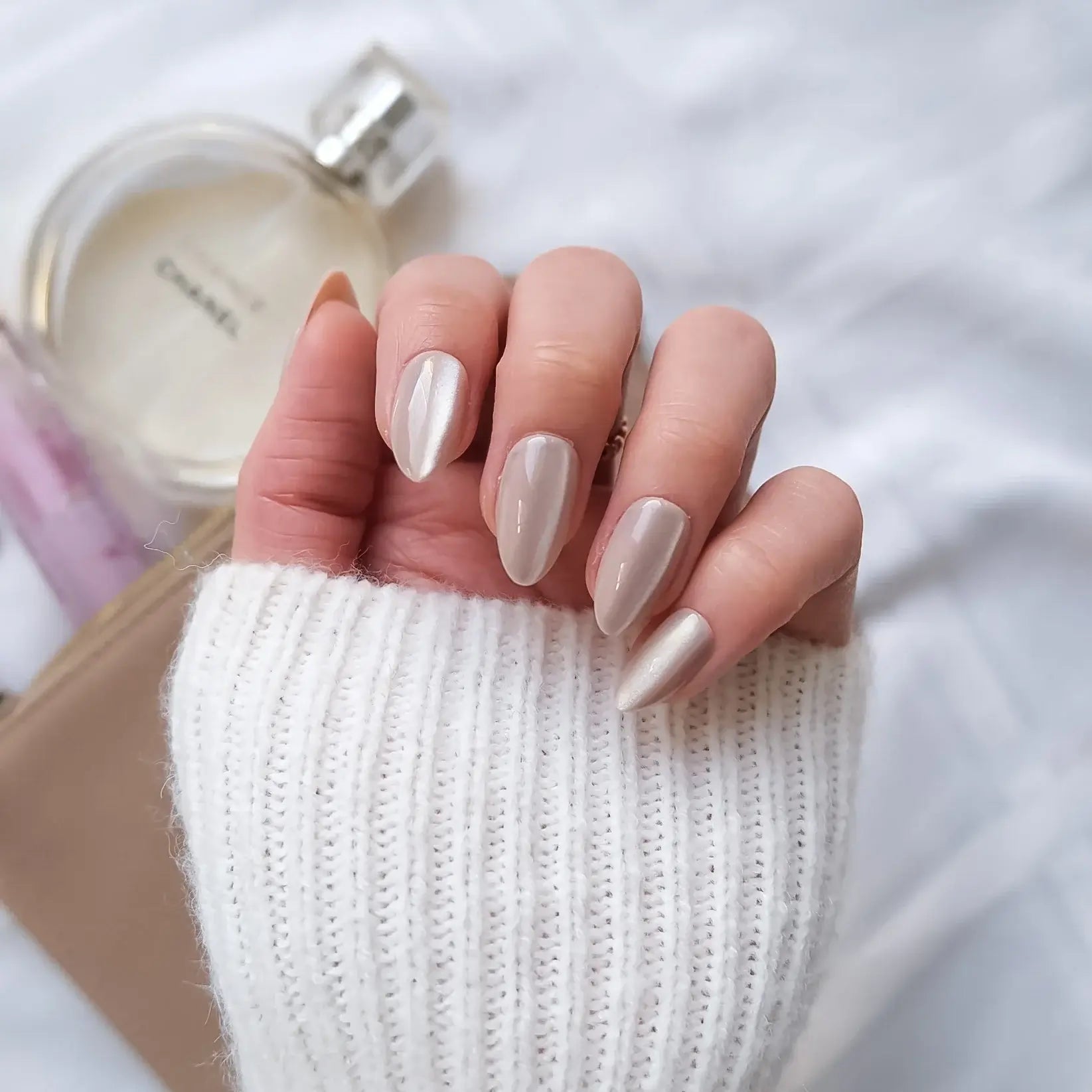 Hand with beige nail press-on nails holding a perfume bottle and phone, wearing a white sweater.